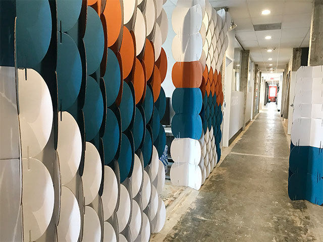 A long room divider made of circular cardboard modules in white, blue, and orange, installed in an office hallway
